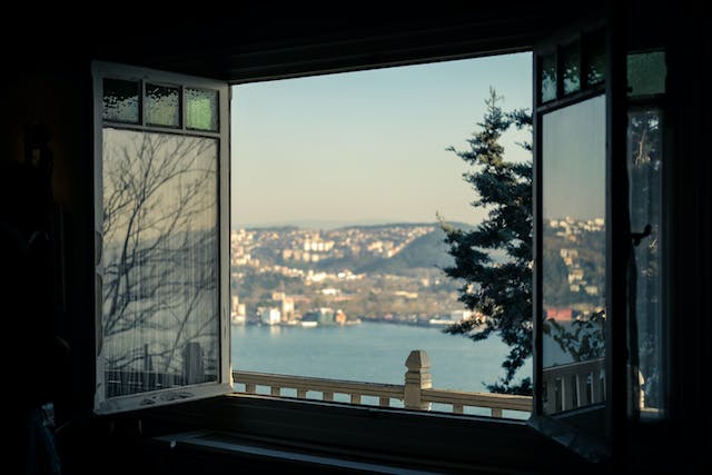 Open bay window showcasing a view of a hilly cityscape and body of water in the distance, with a conifer tree to the right.